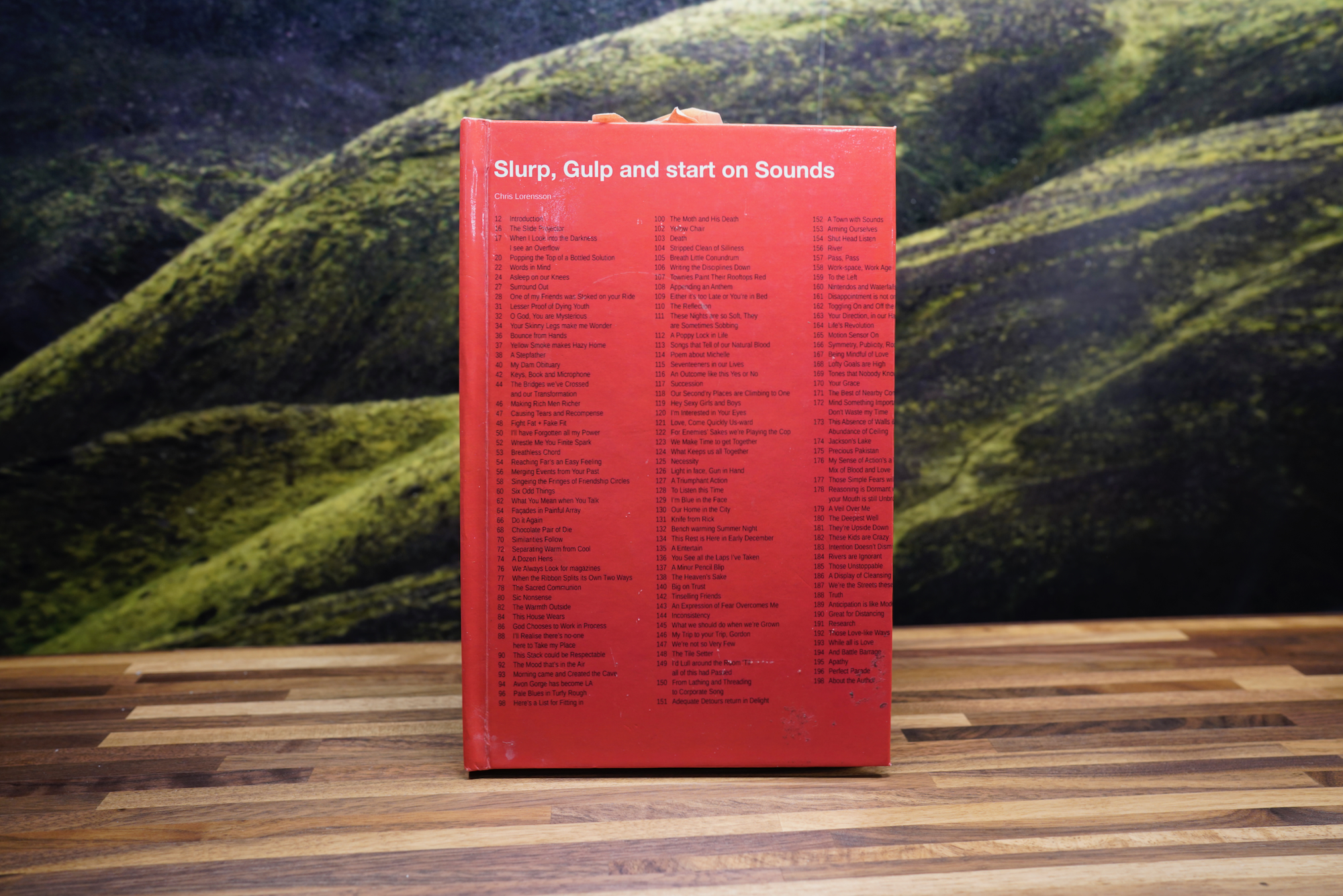 A photograph of the poetry book Slurp, Gulp and Start on Sounds. The book sits atop a butcher-block table with a dark green photophobia of the Icelandic countryside in the near background.